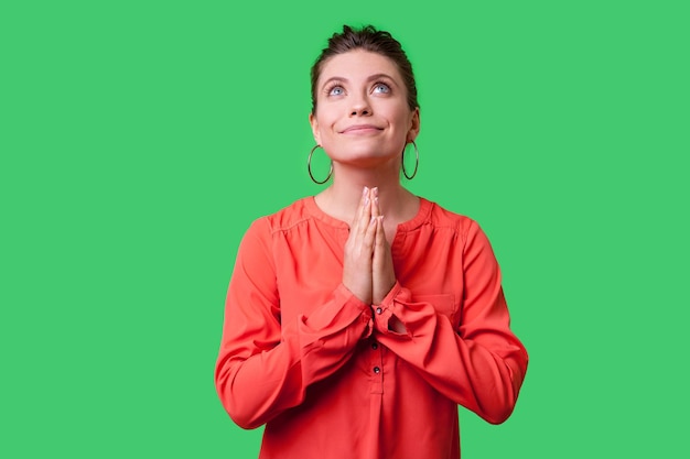 Portret van smekende jonge vrouw met broodje kapsel grote oorbellen en in rode blouse opzoeken met handen in gebed oprechte zeggen gelieve verontschuldiging indoor studio opname geïsoleerd op groene achtergrond