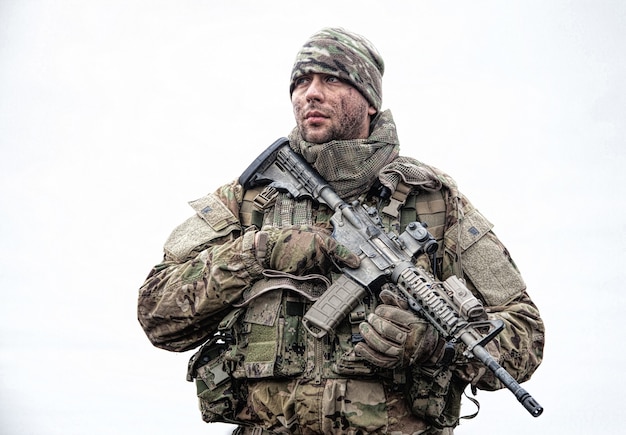 Portret van soldaat, moderne strijder met vuil gezicht, replica van een vuurwapen, camouflage-uniform, muts en maskerende cape op de nek, staande op het veld, rugzak dragend in maart