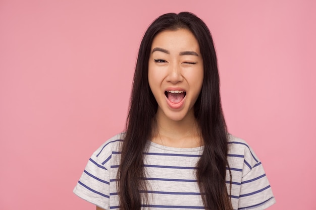 Portret van speels meisje met lang haar knipogend naar de camera en glimlachend in het algemeen enthousiast over kennis met een sluw idee knipperend oog binnen studio opname geïsoleerd op roze achtergrond