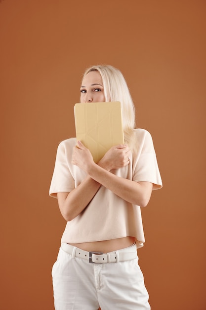 Portret van speelse jonge vrouw met blond haar die mond achter tablet verbergt tegen bruine achtergrond