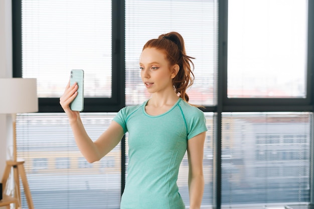 Portret van sportieve vrouwelijke vlogger in activewear die live instructievideo opneemt voor volgers in een lichte kamer op de achtergrond van het raam