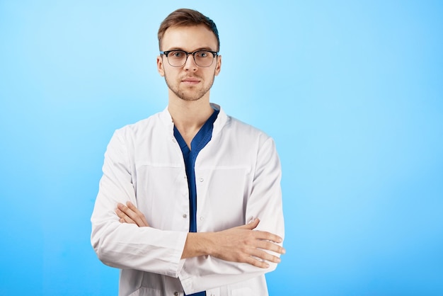 Portret van stagiair arts in witte medische jas en bril met een serieuze gezichtsuitdrukking geïsoleerd op blauwe muur achtergrond