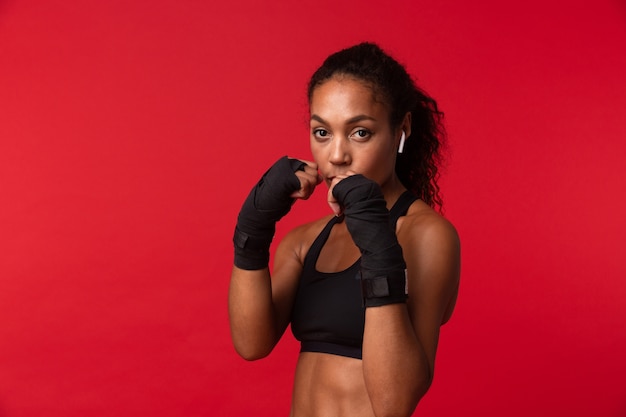 Portret van sterke Afro-Amerikaanse vrouw in zwarte sportkleding boksen met sportverbanden op haar handen, geïsoleerd over rode muur