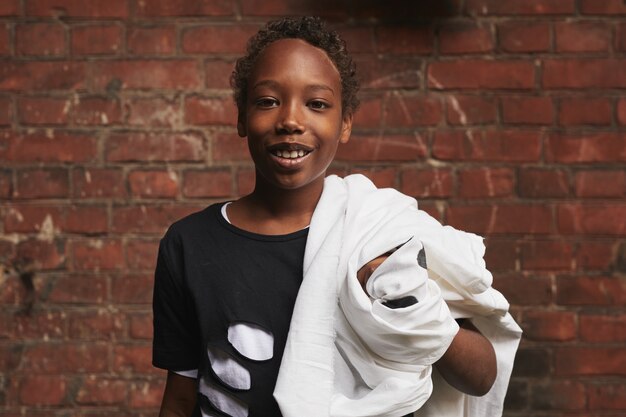 Portret van stijlvolle afro-amerikaanse jongen dragen casual outfit houden spook kostuum staande tegen bakstenen muur glimlachend in de camera