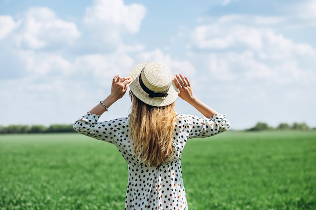 Portret van stijlvolle blonde meisje in hoed op achtergrond van groene lente veld. Een jonge vrouw in een witte jurk loopt in de natuur. Hippie stijl. Groene lente weide. Het meisje houdt een strohoed over haar hoofd.