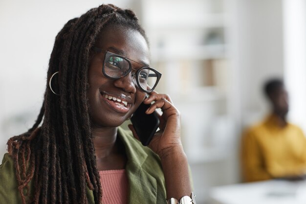 Portret van stijlvolle jonge Afrikaanse vrouw spreken door smartphone in kantoor en glimlachen, kopieer ruimte close-up