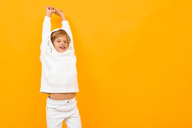 Portret van stijlvolle jonge blanke jongen met half blond haar in witte hoody, zwarte broek geïsoleerd op gele achtergrond