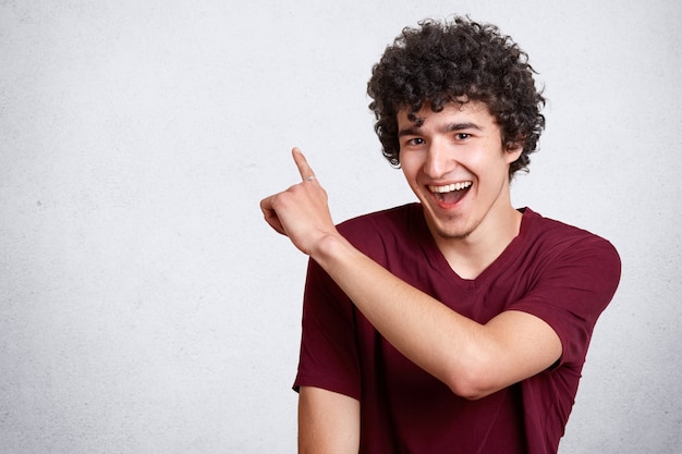 Portret van stijlvolle knappe jonge kerel jurken casual outfit, met donker krullend haar geïsoleerd op wit, wijst een vinger naar de zijkant