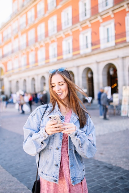 Portret van stijlvolle lachende gelukkige jonge vrouw in de straat met perfecte witte tanden op zoek haar mobiel.