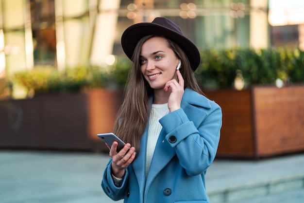 Portret van stijlvolle trendy brunette hipster vrouw in hoed en in een blauwe jas met draadloze witte koptelefoon geniet en luistert muziek in het stadscentrum. Moderne mensen en technologie