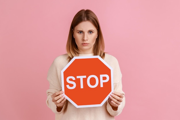 Portret van strikte bazige blonde vrouw met rood stopbord kijken camera met ernstige gezichtsuitdrukking verbod dragen witte trui Indoor studio shot geïsoleerd op roze achtergrond