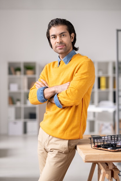 Portret van succesvolle jonge mannelijke manager in gele sweater die zich bij bureau bevinden en wapens op borst in bureau kruisen