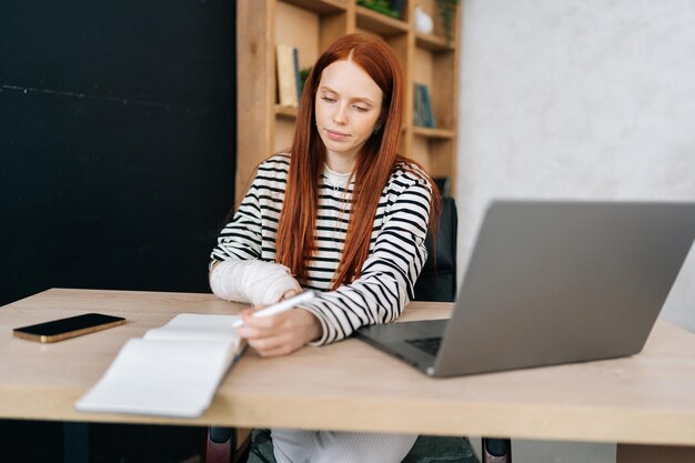 Portret van succesvolle jonge zakenvrouw met gebroken rechterhand gewikkeld in wit gipsverband op afstand werkend met papieren documenten en laptopcomputer
