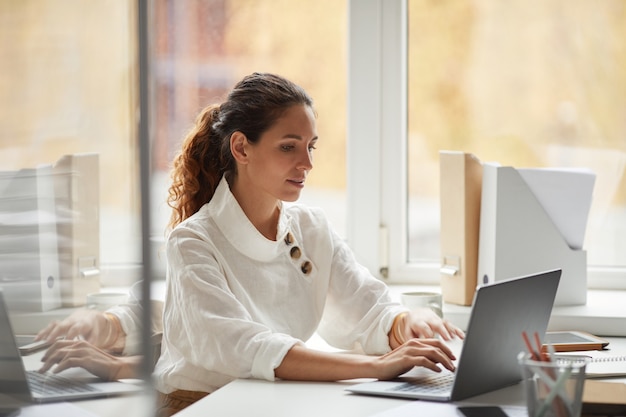 Portret van succesvolle zakenvrouw met behulp van laptop zittend aan een bureau bij raam en genieten van het werk op kantoor, kopieer ruimte
