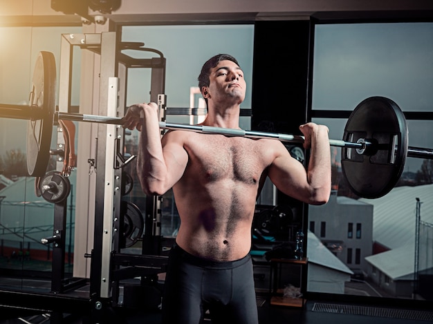 Portret van super fit gespierde jonge man trainen in de sportschool met barbell op grijs