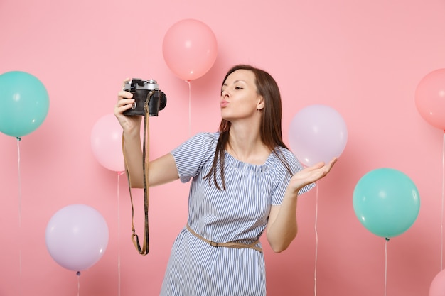 Portret van tedere gelukkige vrouw in blauwe jurk doen selfie op retro vintage fotocamera waait lippen zoenen op roze achtergrond met kleurrijke luchtballonnen. verjaardag vakantie feest mensen oprechte emoties.