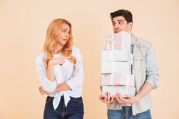 Portret van teleurgestelde vrouw wijzende vinger naar boos man met veel geschenkdozen, geïsoleerd over beige muur