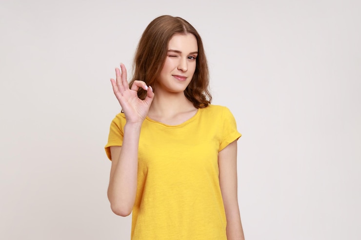 Foto portret van tevreden jonge vrouw met bruin golvend haar in casual gele tshirt staande camera kijken ok teken gebaar en knipogen indoor studio shot geïsoleerd op grijze achtergrond