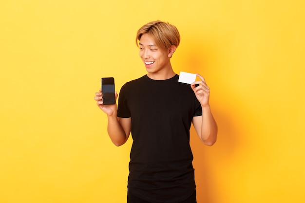 Portret van tevreden knappe Aziatische kerel, die het smartphonescherm en creditcard toont, gsm-scherm, gele muur bekijkt