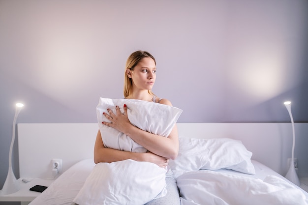 Portret van triest aantrekkelijke vrouw zittend op bed in de slaapkamer en knuffelen kussen