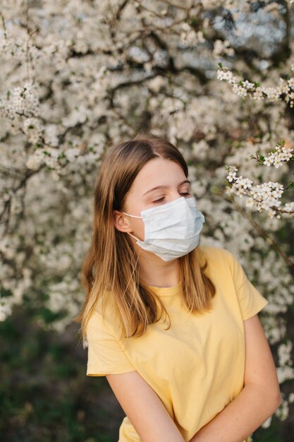 portret van trieste jonge vrouw in beschermende medische gezichtsmasker met bloemen in de buurt van bloeiende boom in het voorjaar. Lente allergie concept