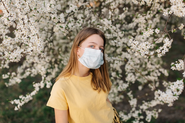 portret van trieste jonge vrouw in beschermende medische gezichtsmasker met bloemen in de buurt van bloeiende boom in het voorjaar. Lente allergie concept