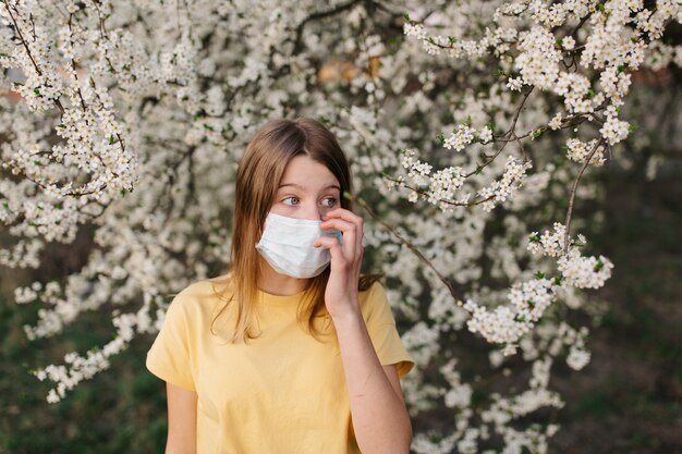 portret van trieste jonge vrouw in beschermende medische gezichtsmasker met bloemen in de buurt van bloeiende boom in het voorjaar. Lente allergie concept