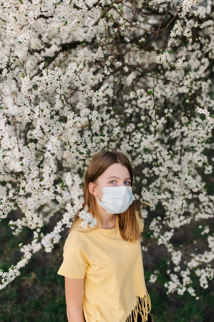 portret van trieste jonge vrouw in beschermende medische gezichtsmasker met bloemen in de buurt van bloeiende boom in het voorjaar.