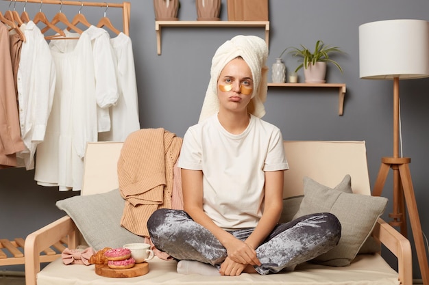 Portret van trieste, overstuur vrouw, gekleed in een witte T-shirt, grijze broek en met een handdoek op het hoofd, in een slecht humeur, uiting van verdriet zittend op hoest met kleren aan hangers op de achtergrond