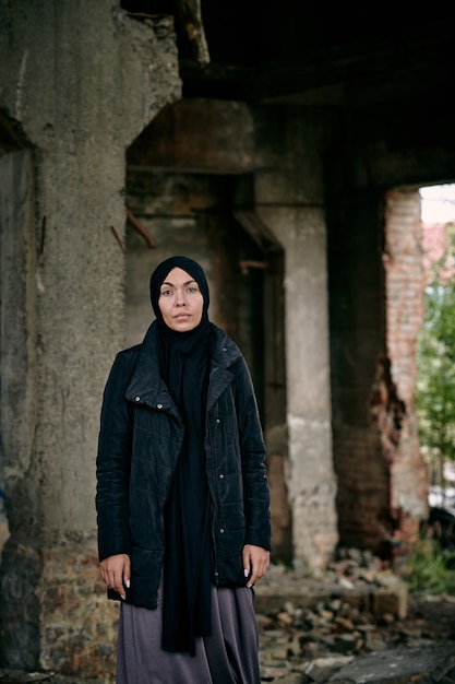 Portret van trieste vluchteling jonge moslimvrouw in hijab en lange jas staande tegen verwoest gebouw