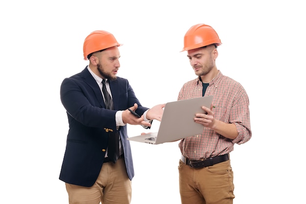 Portret van twee bouwers in beschermende oranje helmen die zich op wit geïsoleerde achtergrond bevinden en laptopvertoning bekijken. Bespreek bouwproject