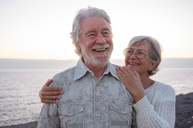 Portret van twee gelukkige en romantische senioren of gepensioneerden omarmd aan de zee bij zonsondergang licht oud lachend senior paar buitenshuis genieten van vakanties samen