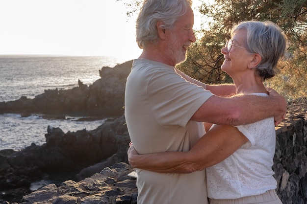 Portret van twee gelukkige en romantische senioren of gepensioneerden omarmd bij de zee bij zonsondergang, lichte oude mensen die buiten samen van vakantie genieten