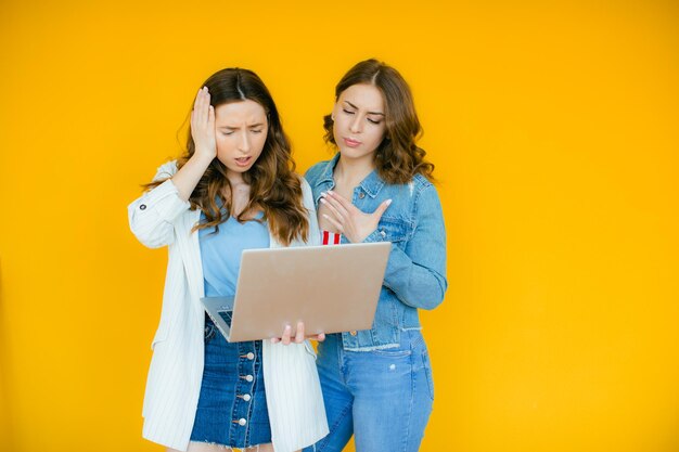 Portret van twee geschokte jonge mee-etermeisjes die met de vinger wijzen op de laptopcomputer op geel