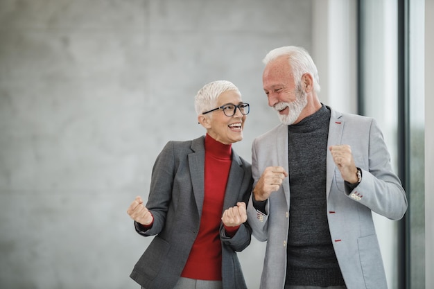 Portret van twee glimlachende succesvolle senior zakenmensen die hun vuisten opheffen terwijl ze in een modern kantoor staan.