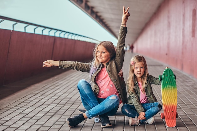 Portret van twee grappige zusjes gekleed in trendy kleding die samen op een skateboard bij een brugvoetgang zitten.