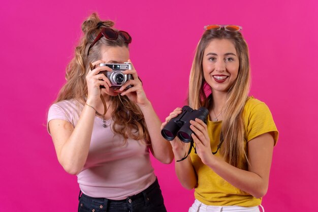 Portret van twee jonge blonde blanke toeristen met camera en verrekijker geïsoleerd op een roze achtergrond