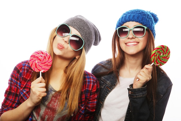 Portret van twee jonge mooie hipstermeisjes die hoeden en zonnebrillen dragen die snoepjes houden Studioportret van twee vrolijke beste vrienden die pret hebben en grappige gezichten maken