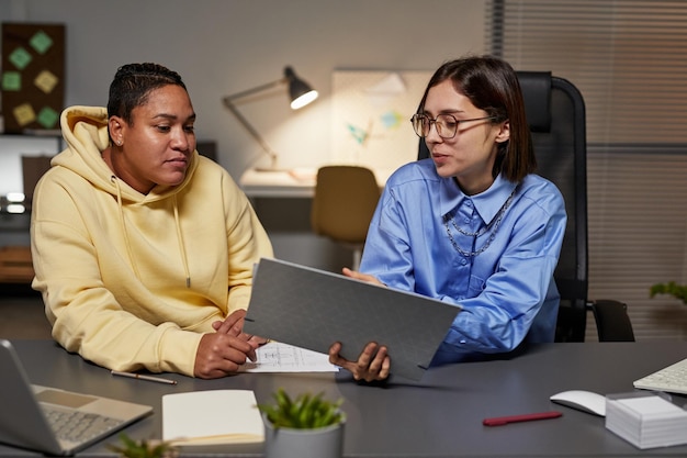 Foto portret van twee jonge vrouwen die documentatie bespreken terwijl ze 's avonds laat op kantoor werken