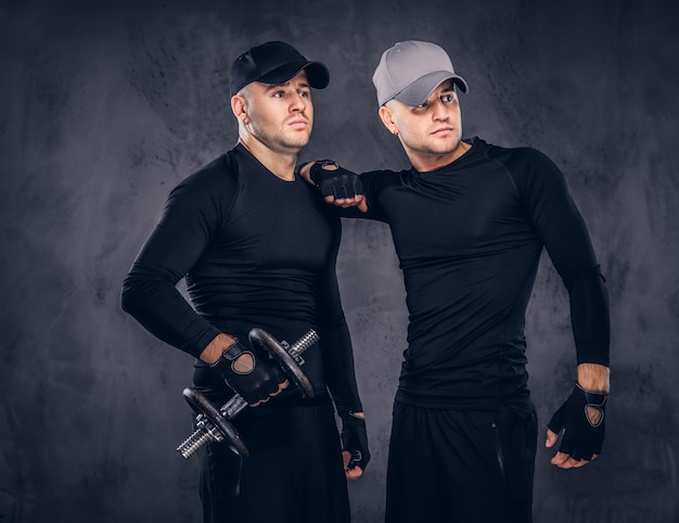 Portret van twee knappe jonge mannen gekleed in een zwarte sportkleding en baseballcap op een donkere achtergrond, houdt een halter vast.