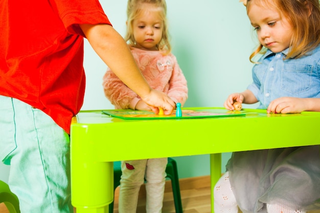 Foto portret van twee lieve meisjes die aan de kindertafel zitten en ongeduldig wachten op het bordspel om te beginnen. twee meisjes zitten van streek na het beëindigen van het bordspel, de jongen verzamelt fiches van de tafel.