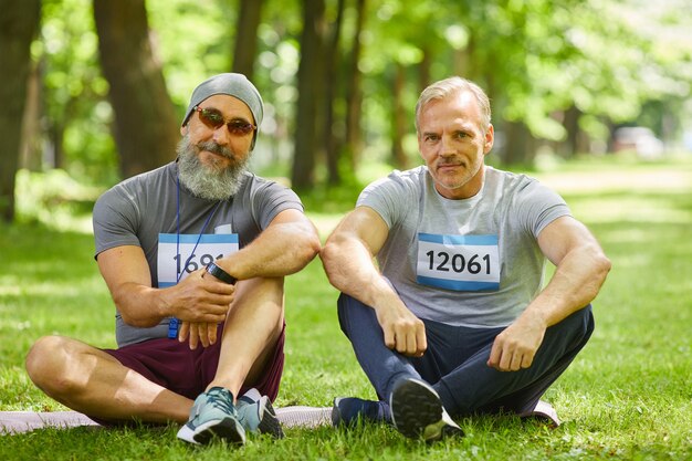 Portret van twee moderne senior sporters die deelnemen aan de zomer marathon zittend op gras in stadspark camera kijken