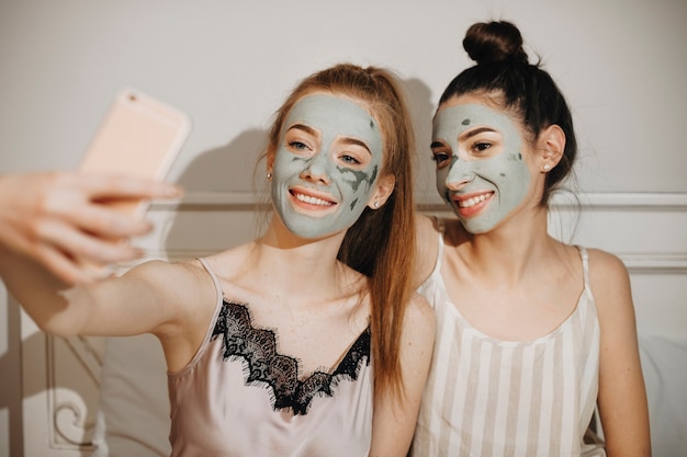 Portret van twee mooie jonge vrouw doet selfie lachen terwijl u ontspant thuis met gezichtsmasker op hun gezicht. Feest voor meisjes die thuis ontspannen.