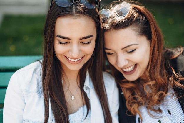 Portret van twee mooie vriendin kijken neer op een smartphone lachen zittend op een bankje buiten close-up.