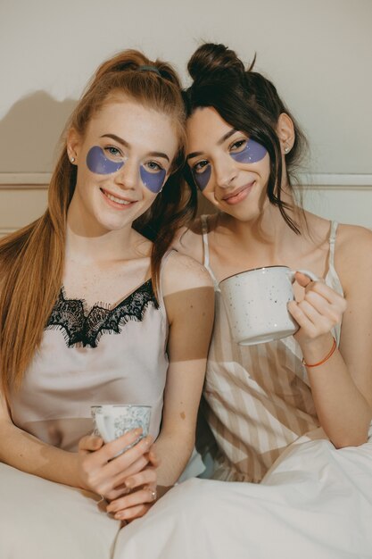 Portret van twee mooie vriendinnen terwijl het drinken van koffie ontspannen op het bed met patches op de ogen.