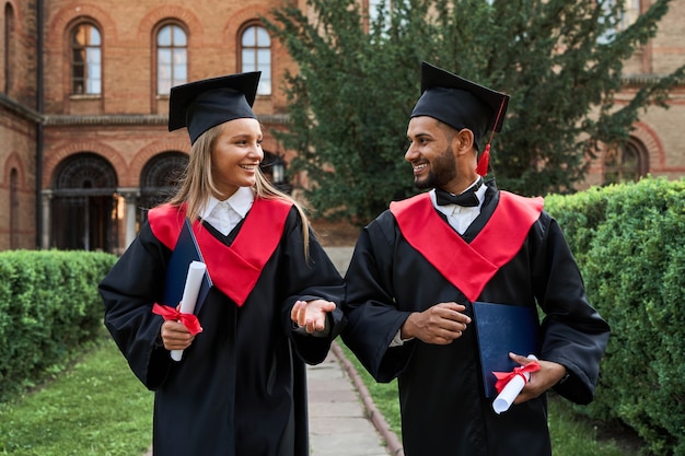 Portret van twee multinationale afgestudeerden die spreken op de universiteitscampus in afstudeergewaden.