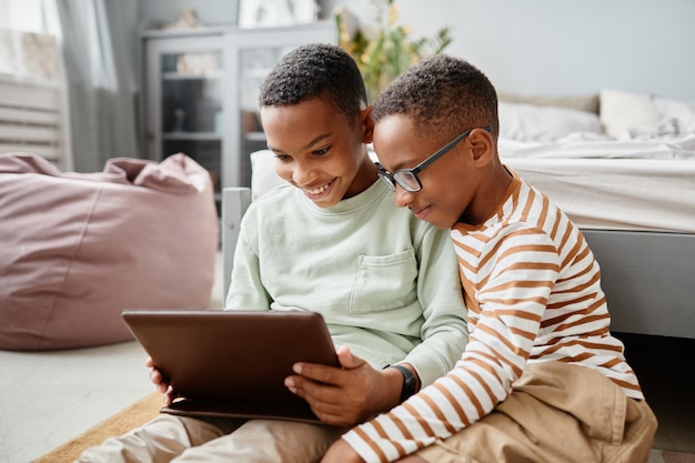 Portret van twee tienerjongens die samen tablet gebruiken terwijl ze thuis op de vloer zitten en video kijken...