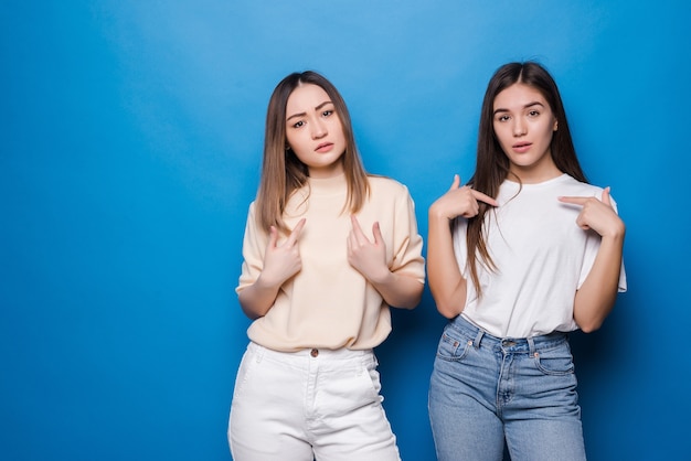 Portret van twee vrolijke jonge vrouwen die zich verenigen en vinger richten zichzelf geïsoleerd over grijze muur