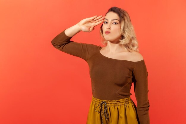 Portret van verantwoordelijke serieuze vrouw met blond haar in bruine blouse groet commandant luistervolgorde met gehoorzame expressie Indoor studio shot geïsoleerd op rode achtergrond