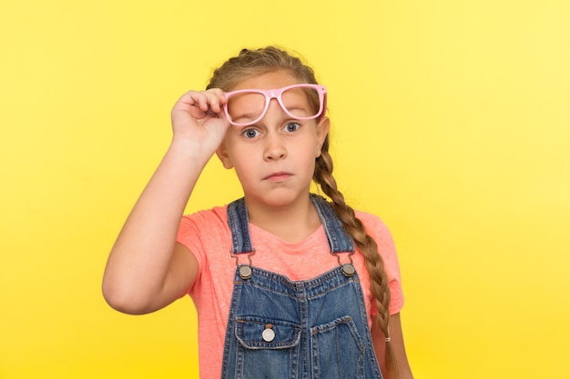 Foto portret van verbaasd stijlvol meisje in denim overalls opstijgen roze bril en kijken met verbaasde uitdrukking kind verrast door haar visie verbetering indoor studio shot geïsoleerd